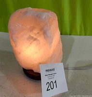 Himalayan white salt lamp glowing on dark wooden base with light on, size approximately 5 x 6.25 x 4 inches visible, photographed with auction number card.