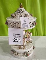 Front view of the white reticulated carousel music box with floral decoration and carousel horses, showing the tag and lot number.