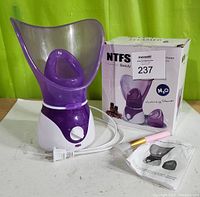 Image of NTFS-618 facial steamer with power cord, instruction manual and original box on white table against green background.