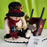 Photo showing the snowman figurine dressed in winter attire shoveling wool/felt snowballs into a decorated red wheelbarrow on a white snow base.