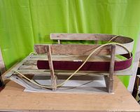 Side view of wooden child sled showing the slatted seat, curved front with maroon reinforcement, and rope handle.