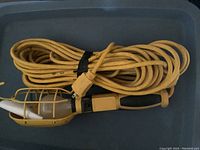 Photo shows a DeWalt branded handheld utility trouble light with a protective yellow metal cage around the bulb and a yellow and black handle. Coiled neatly beside the light is a long yellow electrical cord approximately 50 feet in length.