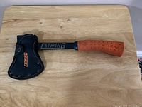 Full view of Estwing hatchet with sheath over the blade, showing orange rubber grip handle and wood-grain top surface.