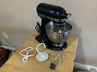 Black KitchenAid 10-speed electric stand mixer with stainless steel bowl and attachments (flat beater, dough hook, wire whip) on a wooden table.