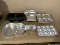 Front angled view of all five bakeware pieces on the floor showing guitar cake pan, black double muffin pan, heart-shaped tiered cake pan, and two aluminum baking molds.