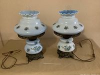 Pair of white glass hurricane lamps with floral designs and ornate metal bases, placed side by side