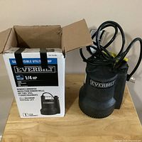 Photo of Everbilt submersible utility pump placed next to its original packaging box on a wooden surface, showing brand and key features on box.