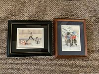 Two framed prints on carpeted floor. Left is a hockey scene titled 'Tripped Up' by John Newby, right is 'Taking a Breather' by Shirley Deaville.