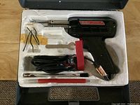 Open case showing Weller Expert soldering gun pistol grip tool with metal tip, multiple soldering tips, small wrench, wires, and red cleaning tool arranged in fitted white foam insert.