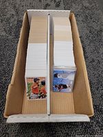 Box containing two stacks of baseball cards, one stack with cards facing down and visible blank backs, the other with top card featuring a young baseball player with a bat.