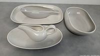 Top-down photo showing gravy jug on oval plate, large bowl to right, and rectangular serving plate on left, all light gray ceramic.