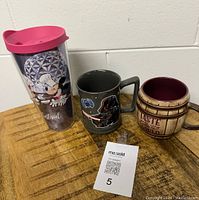 Set of three coffee mugs including Minnie Mouse Tervis tumbler, Darth Vader Star Wars mug, and barrel-style mug displayed on wood surface.