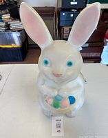 Front angled view showing full figure of the white blow mold Easter bunny with pink ears and nose, blue eyes, holding a basket with colorful eggs.