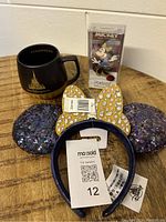 Photo showing black Starbucks mug with golden Disney 50th anniversary castle design, Minnie Mouse sequin ears with gold bow and white jewels, and Mickey Figpin in package