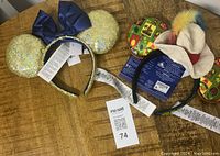 Two Disney 50th ears headbands on wooden surface, one gold glitter with blue bow, one colorful patch design with hat and feathers, all with tags.