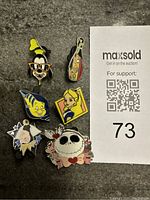 Photo of six Disney enamel trading pins laid out on a dark surface next to a MaxSold auction paper.