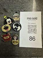 Photo of 6 Disney trading pins arranged on a gray surface with a maxsold auction support card next to them.