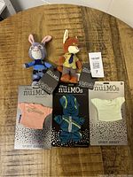 Full overview of the two Disney nuiMOs plush toys and three pieces of plush clothing with original packaging and tags on a wooden surface.