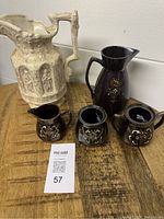 Front view of the large cream-colored embossed jug alongside three smaller dark brown redware pottery pieces with hand-painted floral decorations placed on a wooden surface.