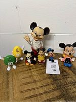 All items shown grouped on wood table surface, includes large Mickey and Minnie Mouse figurines, several smaller Disney figures, and two M&M candy dispensers in green and yellow.