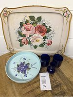 Photo showing two rectangular vintage tin trays with floral rose designs behind blue glass items on a wooden surface.