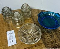 Lot of three vintage glass insulators and two ashtrays on wooden table. One of the insulators shows visible damage. Included blue circular glass ashtray and a clear ashtray with concentric rings.