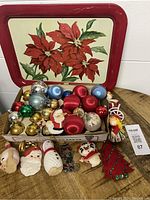 Wide view showing the poinsettia tin tray with assorted colorful vintage Christmas ornaments including ball-shaped and character figurines spread out on the wooden surface and in a box.