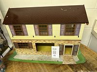 Front view of the vintage tin metal dollhouse showing detailed exterior including windows with purple shutters, brick and yellow siding, and damaged roof spots.