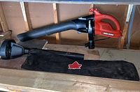 Black & Decker Vortex electric blower assembled with a straight nozzle, laying on a wooden surface next to a black storage bag with red maple leaf logo and an additional nozzle attachment.