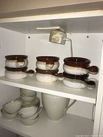 Six ceramic French onion soup bowls with built-in handles on a shelf, paired with matching underplates in dark brown and speckled beige.