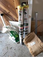 Photo of two rolled weed block sheets labeled Need Block, partially used turf fertilizer bag, burlap sheet folded on the floor, and a metal ruler leaning against wall.