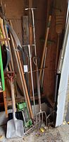 Garden tools including shovels, rakes, fork, and pruning saw leaning in corner of garage.