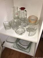Shelf with assorted glassware: pitchers, bowls, covered dish, serving trays including a ceramic bowl and clear glass items