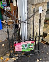 Black metal gate panel leaning against garage wall outdoors, showing overall design and dimensions with a plastic bag fixed to it.