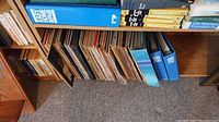 Long view of the collection of LP vinyl records arranged on a wooden shelf beneath other books and binders, showing many record spines varying in color and thickness.