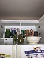 Wide shot of cabinet shelf showing a variety of pottery and glass vases including a white ceramic bowl with handles at the front right, and several colorful vases behind.