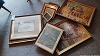 View of multiple framed prints arranged on carpet, showing variety of sizes and frame styles, including gold and wood frames