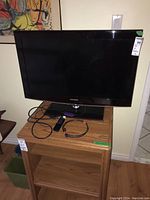 Full view of Samsung 32 inch TV showing front screen and base stand on wooden furniture with cords and remote visible.