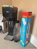 Black SodaStream sparkling water maker, clear Source bottle with black cap, original boxed spare CO2 carbonator canister positioned on carpet near beige wall.