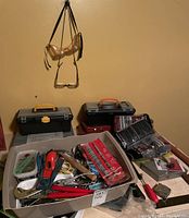 Box containing assorted hand tools in various conditions located on table with two black toolboxes behind and safety goggles hanging on wall.