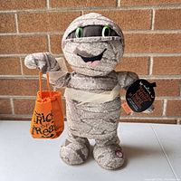 Front view of the Halloween Dancing Trick or Treat Mummy plush holding orange trick-or-treat bag