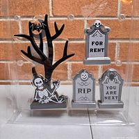 Front view of four Halloween mantle decor figures in plastic packaging showing details including a black tree with skeleton and bats, and three tombstones with messages
