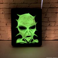Front view of the glowing 3D alien wall decor illuminated in green light, showing detailed facial textures and alien head spikes.
