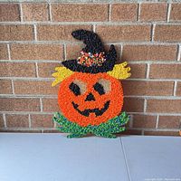 Front view of vintage Halloween plastic popcorn pumpkin wall hanging with a smiling face, witch hat, yellow hair, and green leaves at bottom, displayed against brick wall.