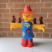 Front view of scarecrow target toy showing face, red hat, blue vest with bullseye, red pants, and black/red metal hinged targets on both arms.