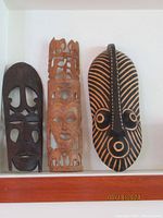 Three vintage African wood tribal masks displayed side by side. Left mask dark brown with cut outs, middle mask tall natural wood with carvings of birds and elephants, right mask oval with black and natural stripes and circular carved features.