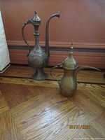 Pair of antique vessels on wooden floor against brown wall: taller tinned copper water ewer with curved spout and handle; shorter vintage brass Arabic coffee pot with bent spout and finial lid.