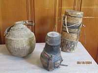Three antique African milk jugs: two rounded with fiber covering and leather straps, one cylindrical wooden with dot pattern and leather straps