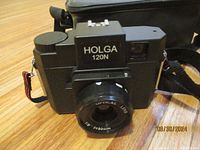 Holga 120N medium format film camera on wooden surface with carrying case behind.