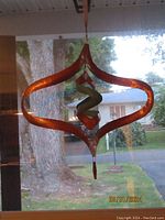 Copper wind spinner hanging indoors against a window background, showing its diamond-shaped frame and inner spiral design.